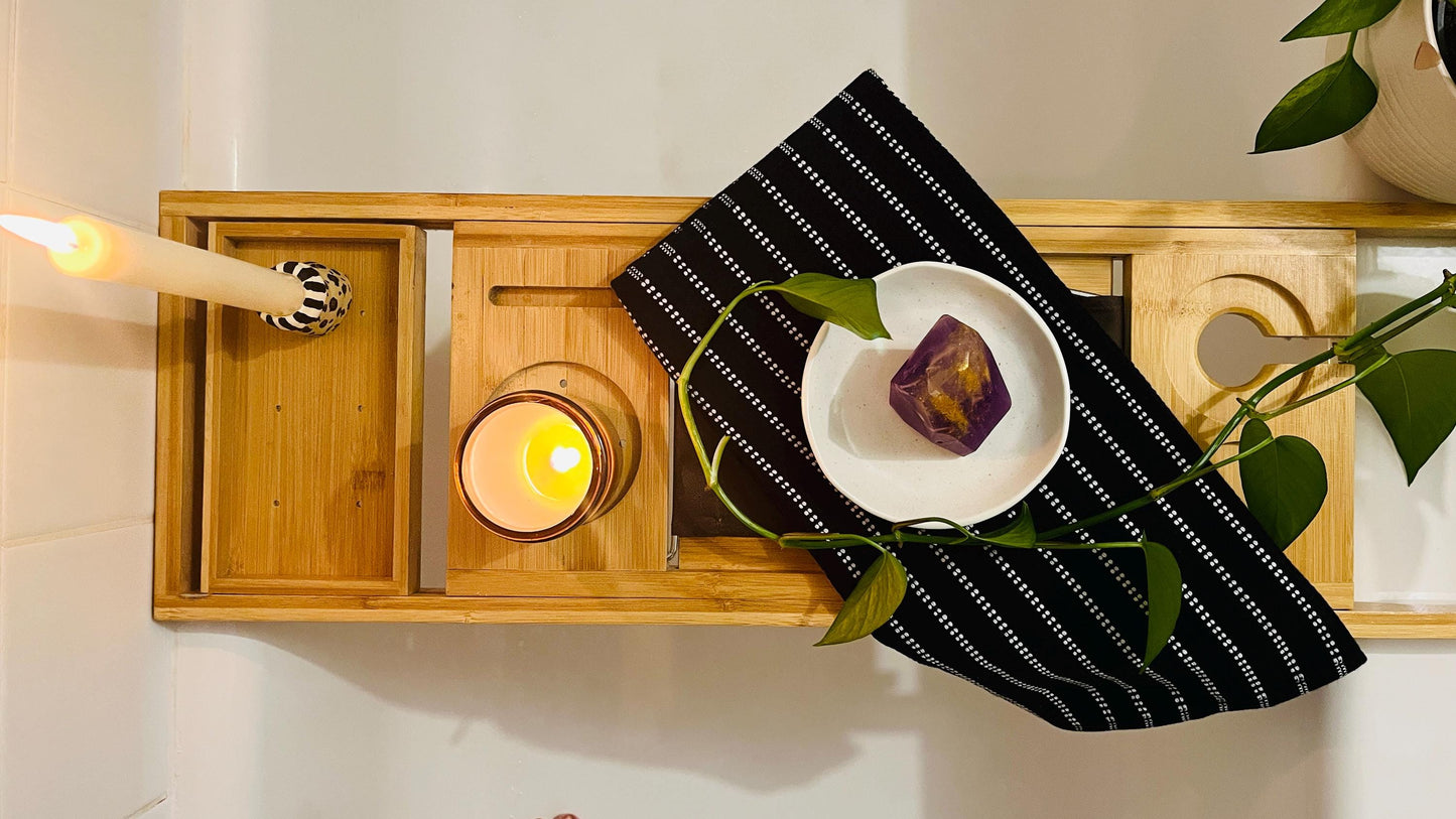 Wooden Bathtub tray with candle, purple gem shaped soap, and black and white towel with plant beside it
