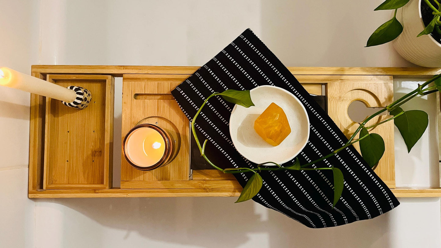 Wooden bathtub tray with candle, orange gem shaped soap, black and white striped towel, and plant on a bathtub ledge.