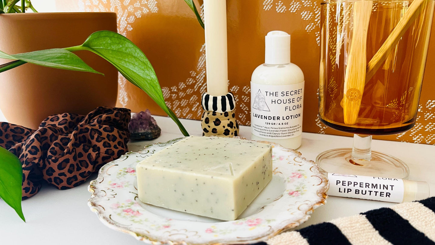 Cosmetic products including a bar of light green soap, lotion bottle, and lip balm on a decorative vintage soap dish with a plant and candle in the background.