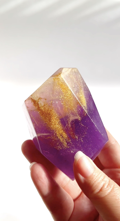 Hand holding a purple and gold gem shaped soap against a white background