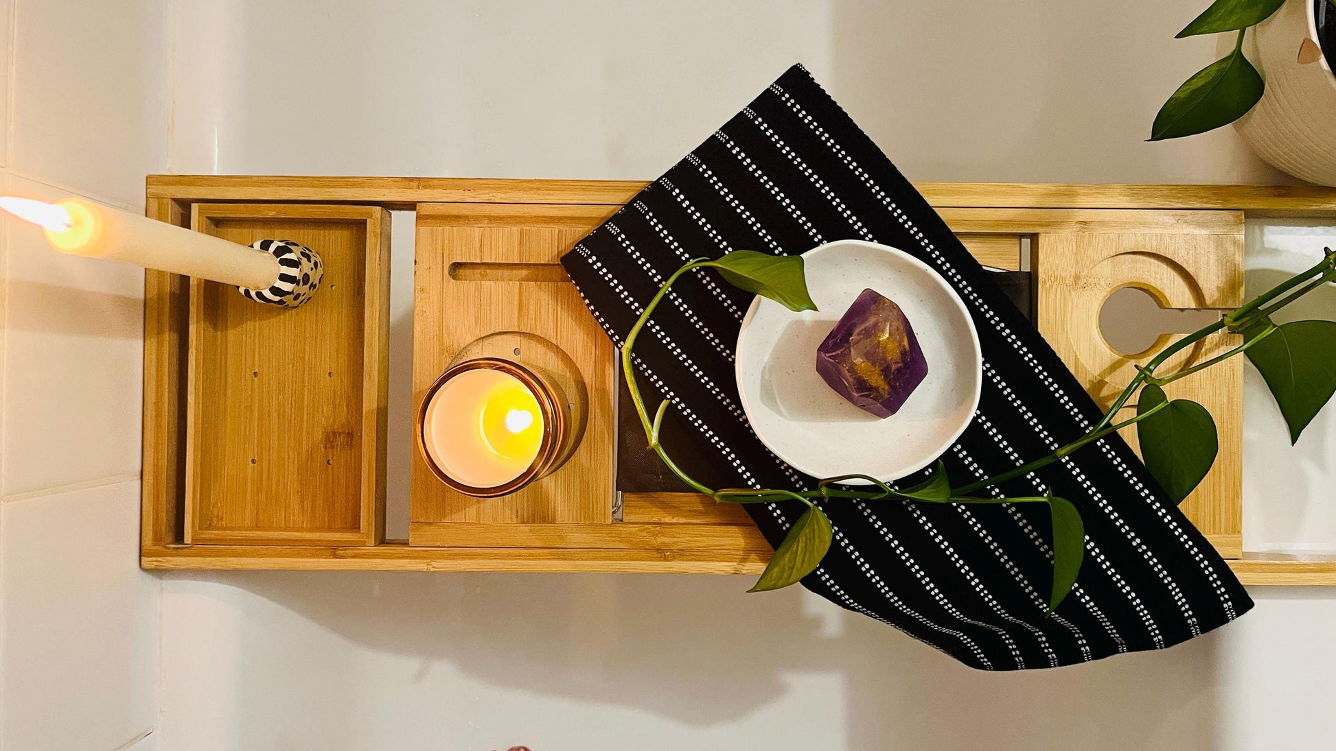 Wooden Bathtub tray with candle, purple gem shaped soap, and black and white towel with plant beside it
