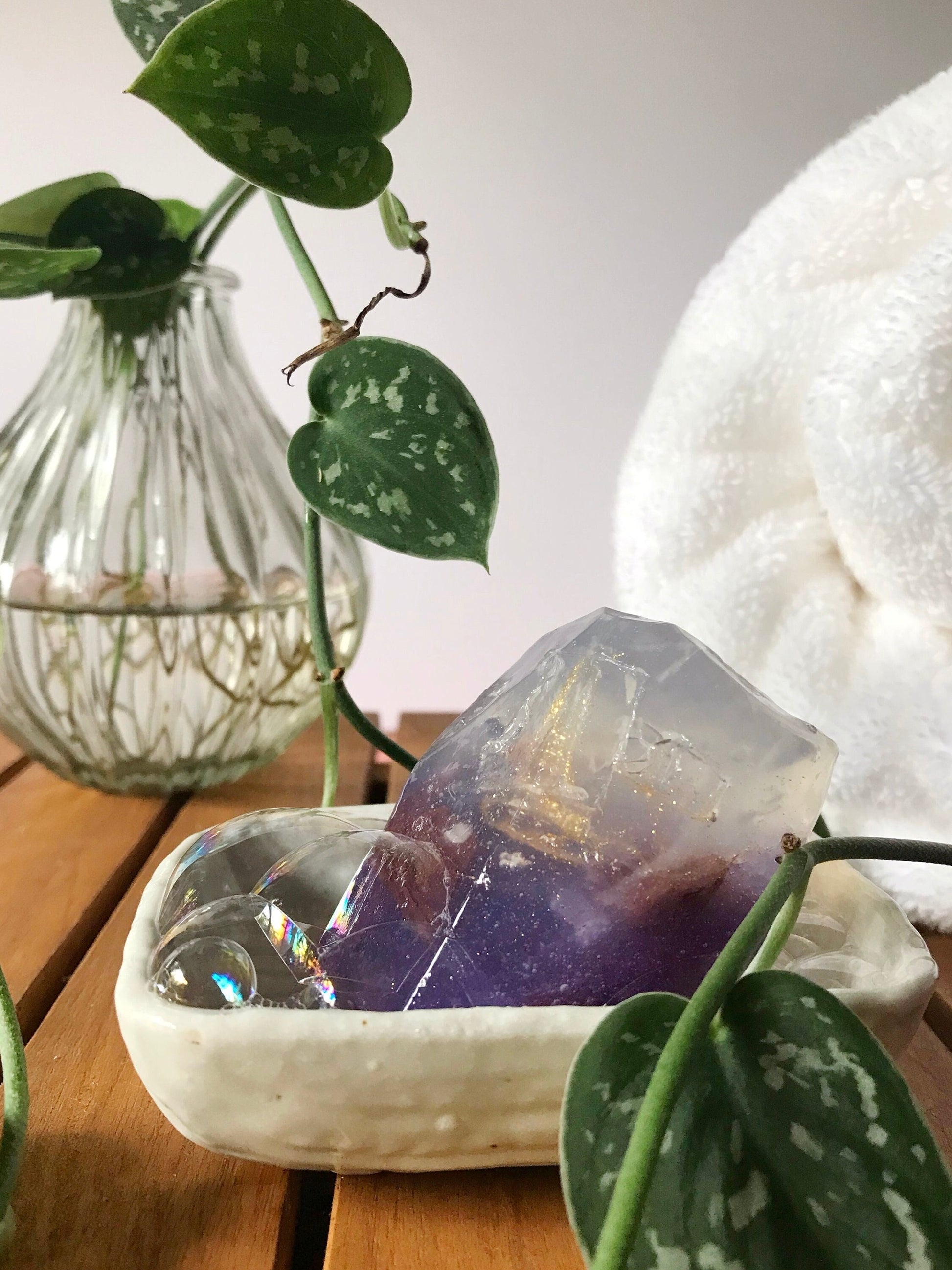 Purple gem shaped soap in a white dish with plants, and a white rolled up fluffy towel in the back ground.