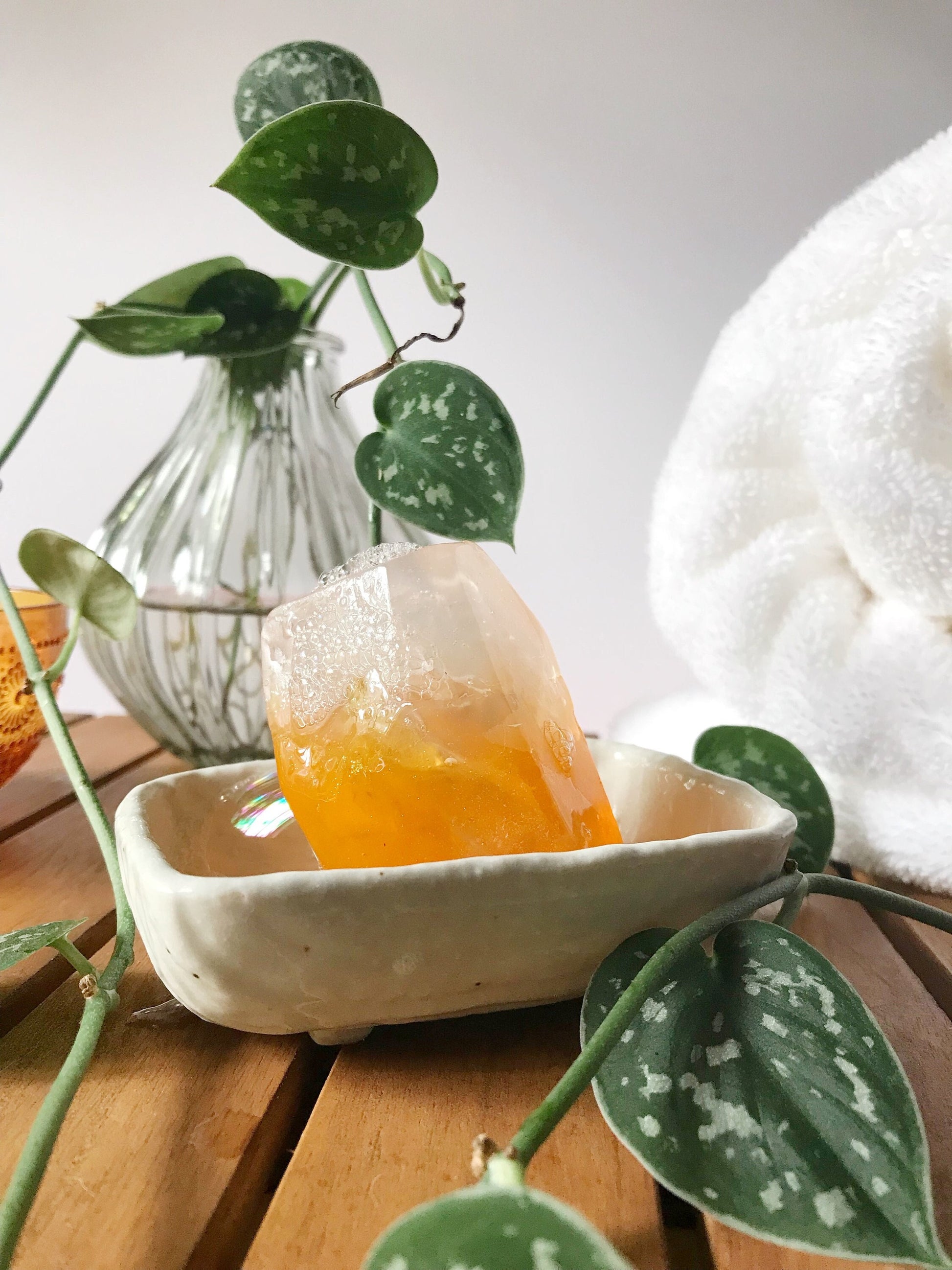 Orange gem shaped soap in a white soap dish with plant and fluffy white towel in background.