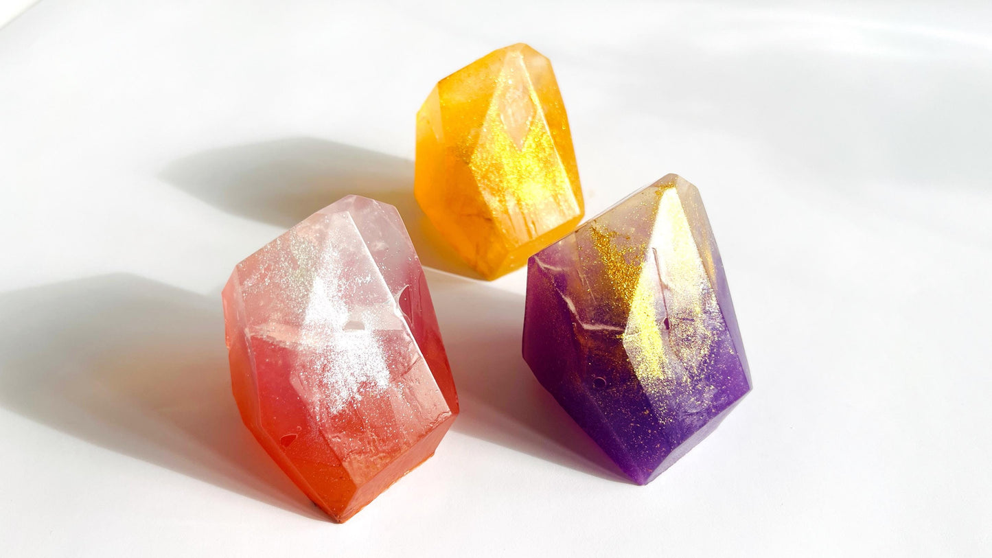 Three colorful crystal gen shaped soaps (rose quartz, citrine, amethyst) on a white background