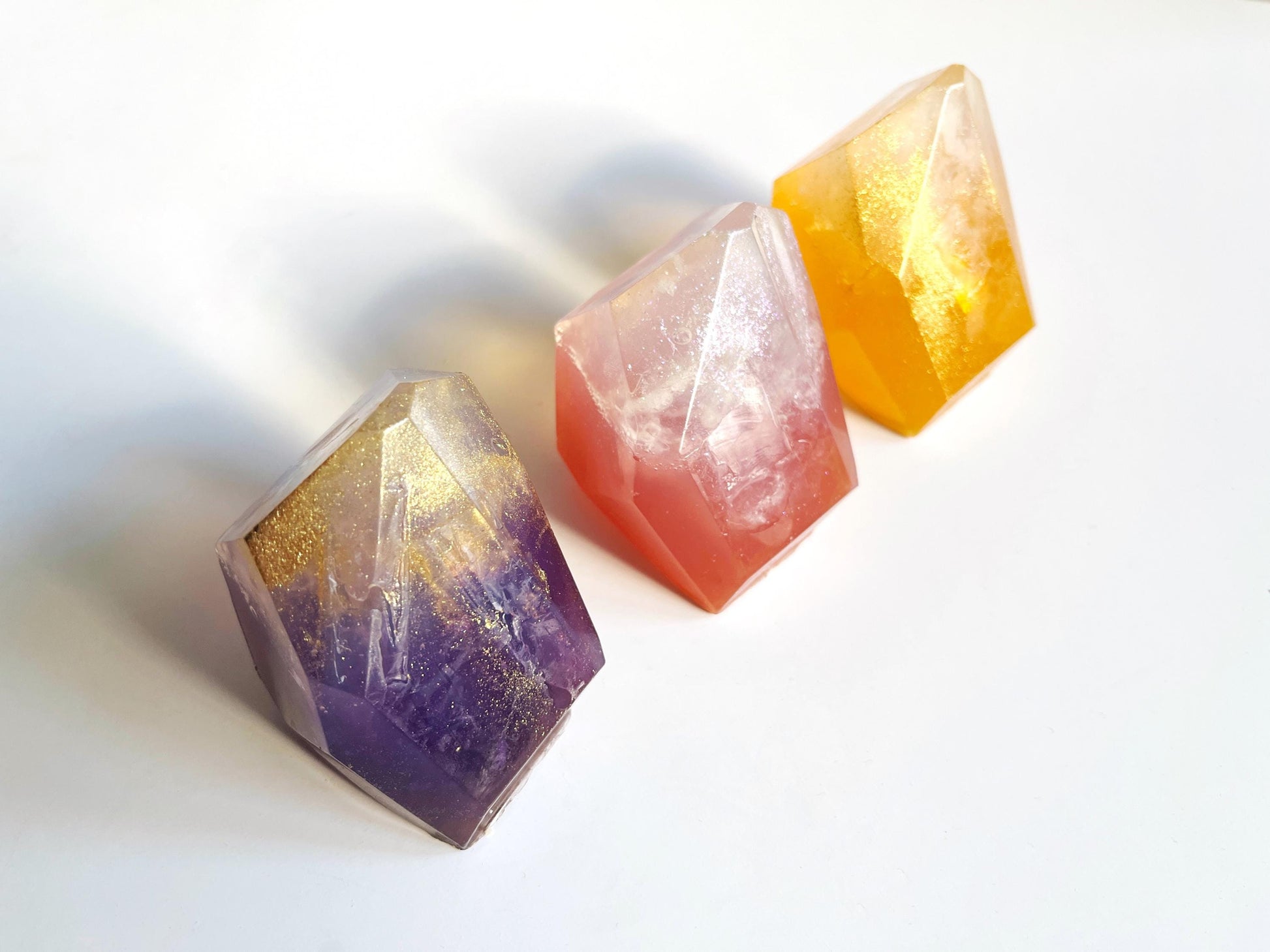 Three crystal-like gem shaped soaps in purple, pink, and orange on a white background