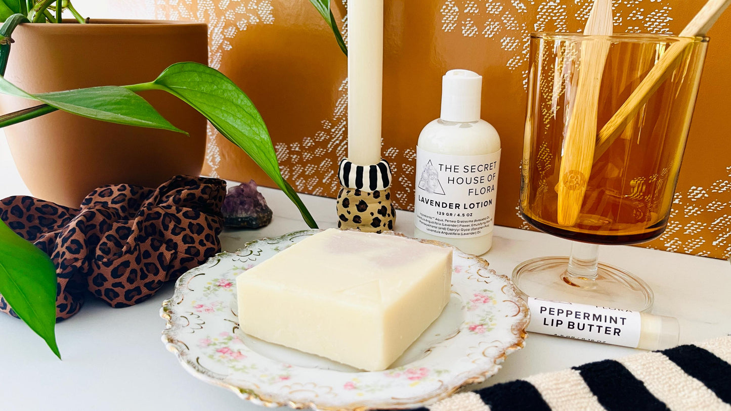 Bar of soap on a decorative vintage floral  soap dish with a bottle of lotion, candle, and other items on a table.