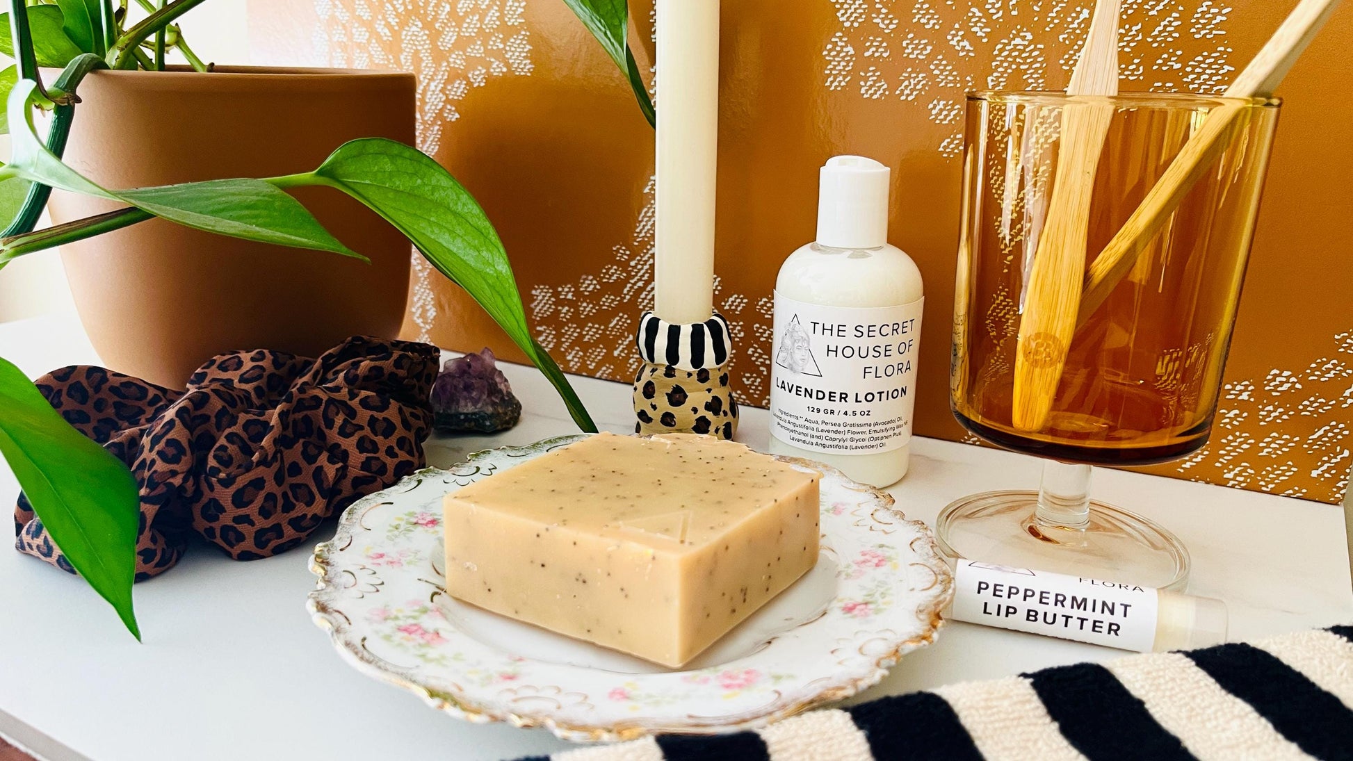 Cosmetic products including a bar of light yellow soap, lotion bottle, and lip butter on a decorative vintage floral soap dish with a candle and plant in the background.