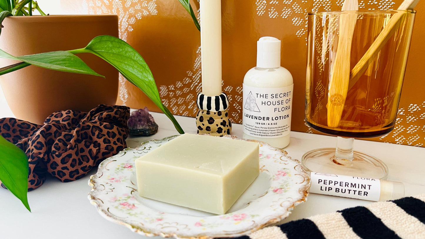 Bar of  sage green soap on a vintage floral soap dish with a bottle of lotion, jar, and other items on a table.
