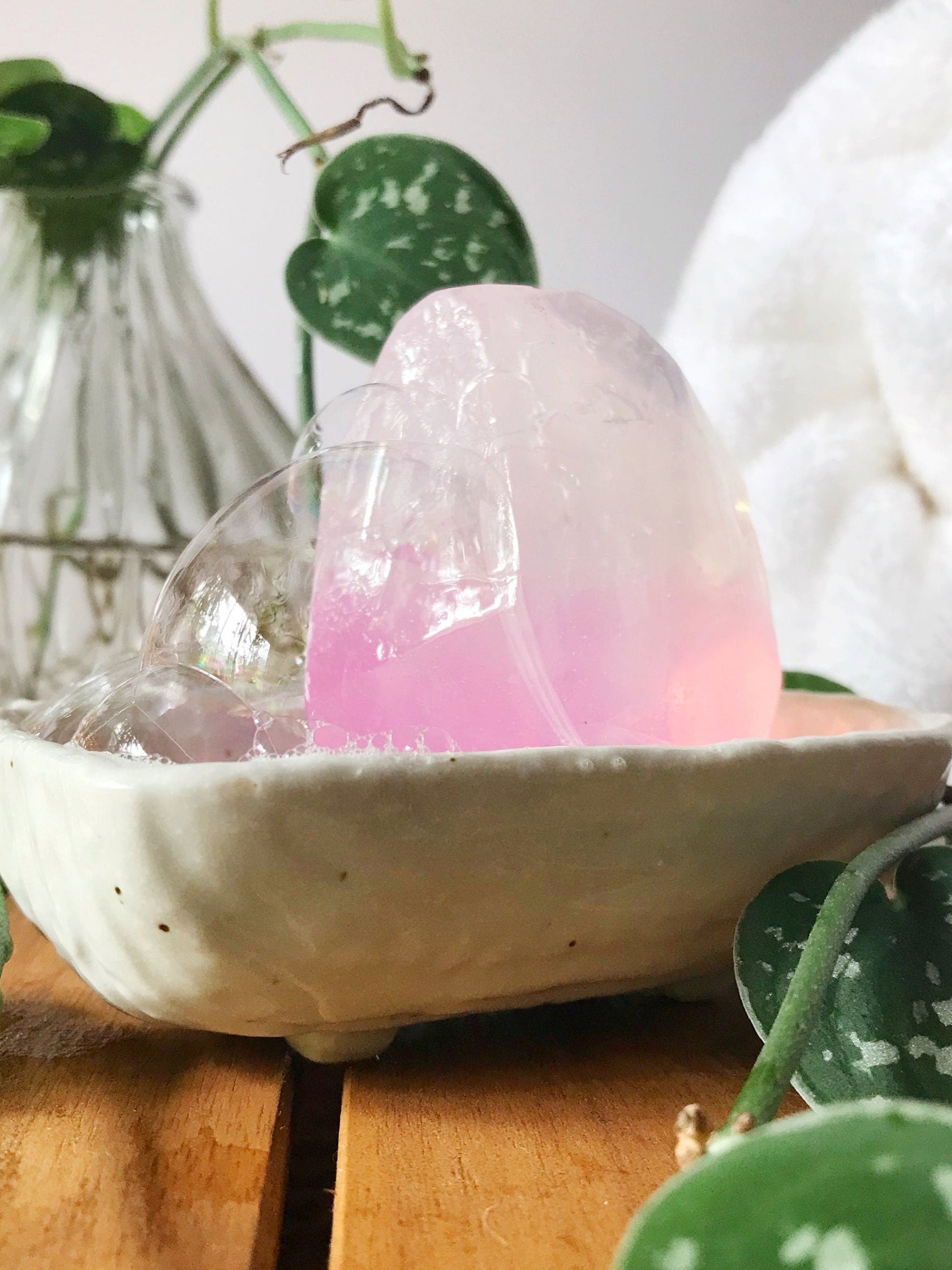 Pink gem shaped soap in a white dish with green plant, and a white fluffy rolled up towel in the background.