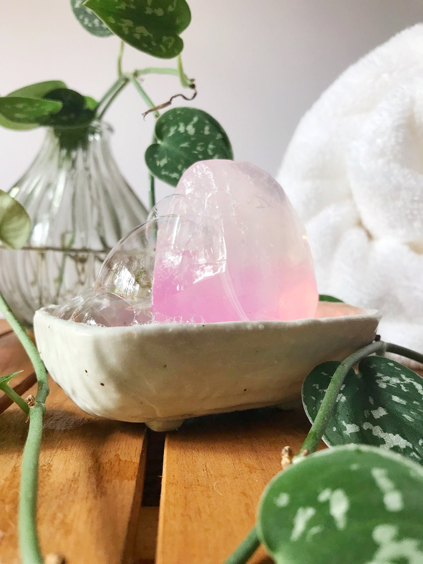 Pink gem shaped soap in a white dish with green plant, and a white fluffy rolled up towel  