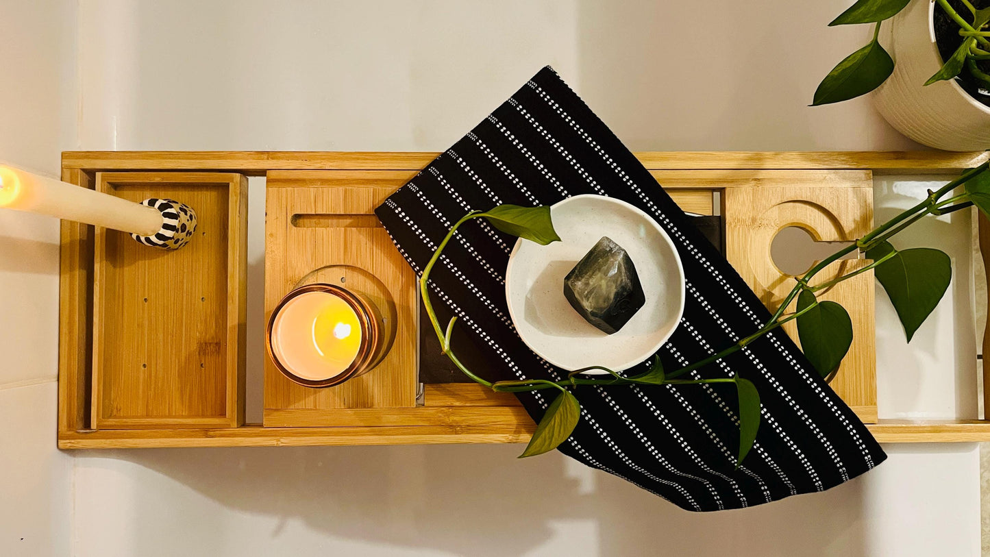 Wooden Bathtub tray with candle, smoky black gem shaped soap, and black and white towel with plant beside it