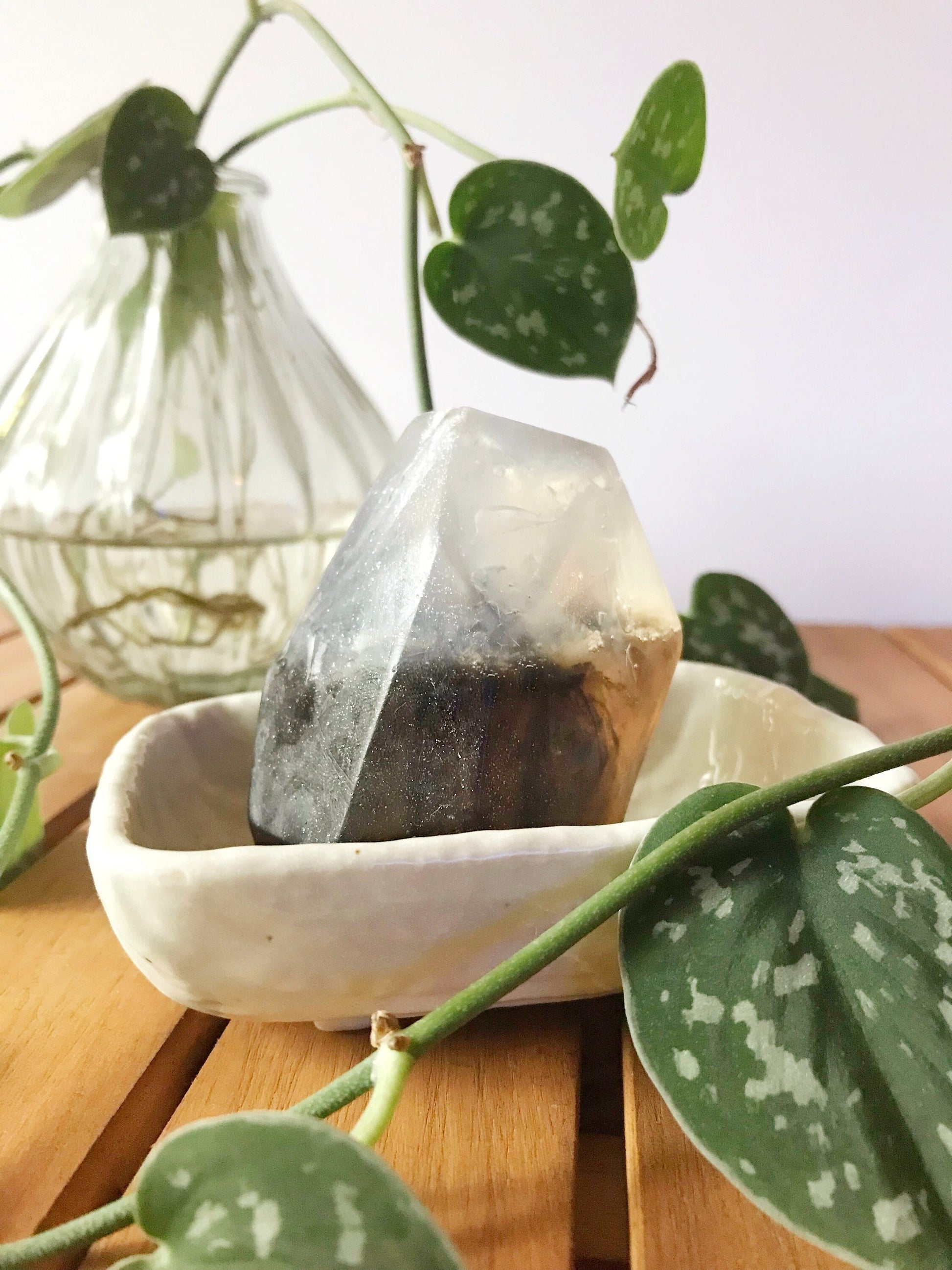 Smoky black gem shaped soap in a white soap dish on a wooden surface with plants in the background.