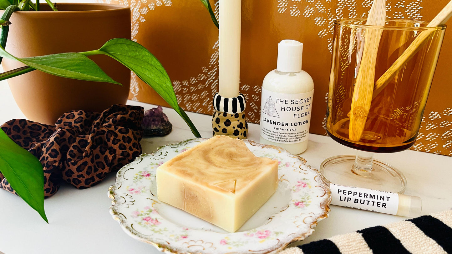 Bar of orange and white marbled soap on a decorative vintage floral soap dish with a bottle of lotion, tube of lip butter, and other items on a table.