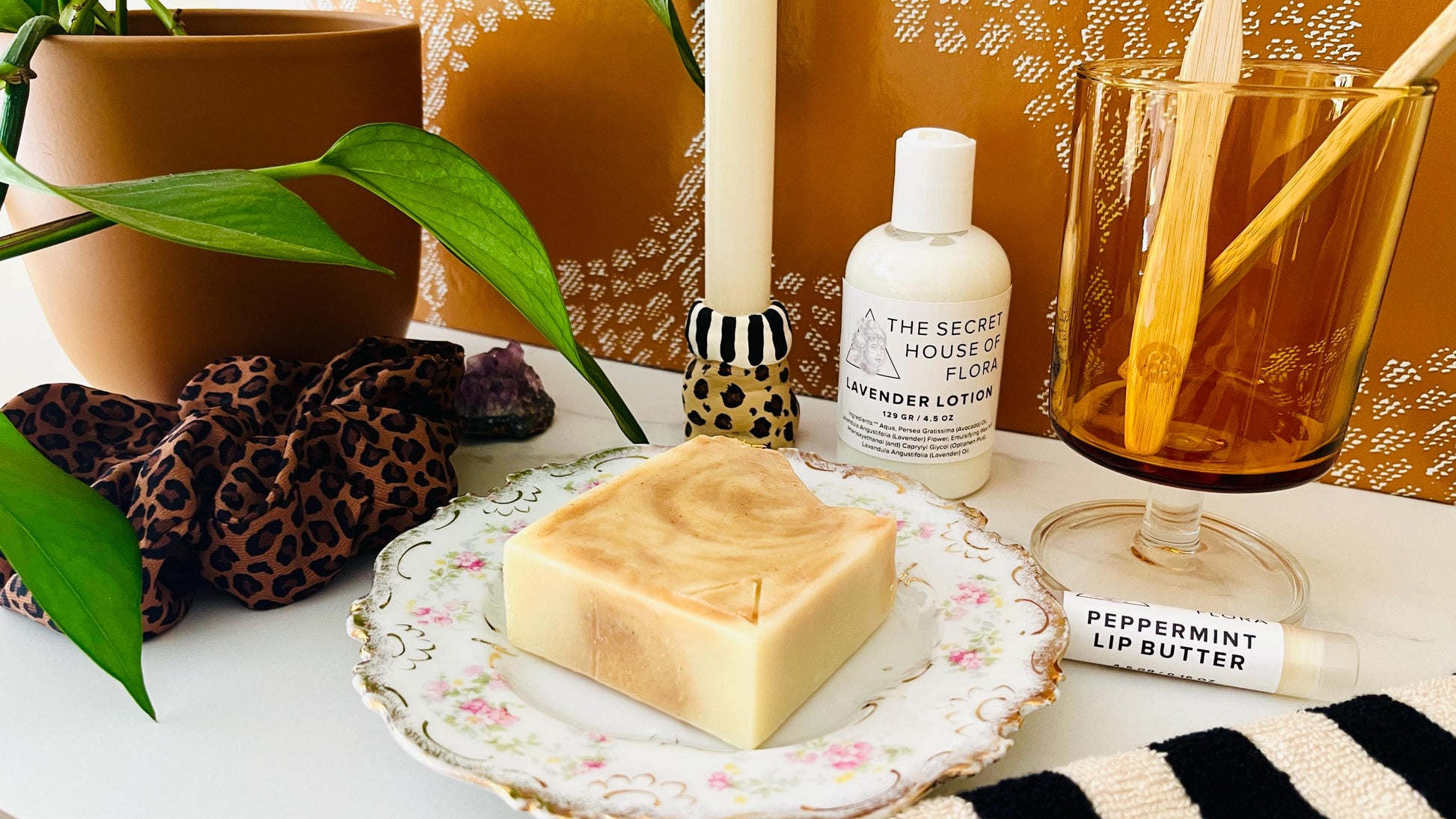Bar of orange and white marbled soap on a decorative vintage floral soap dish with a bottle of lotion, tube of lip butter, and other items on a table.