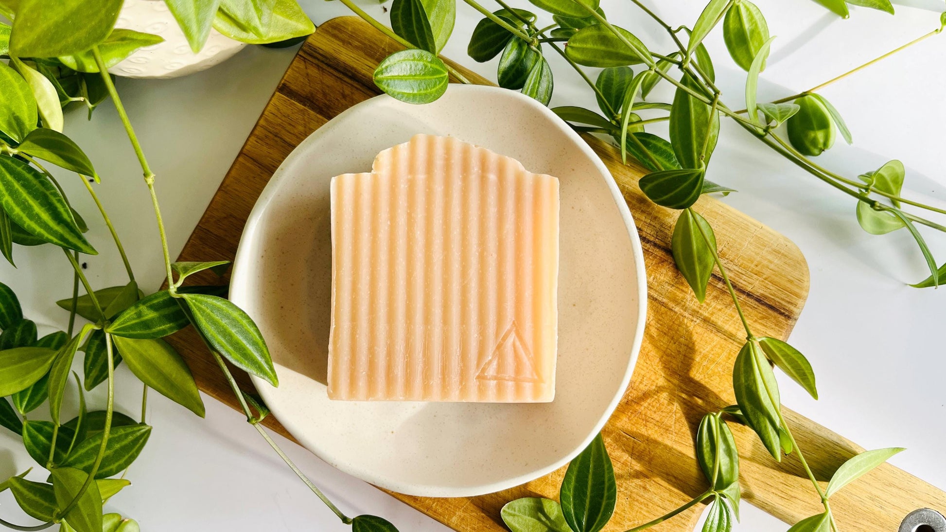 Bar of pink soap with soft ridges on a white soap dish with green leaves around.